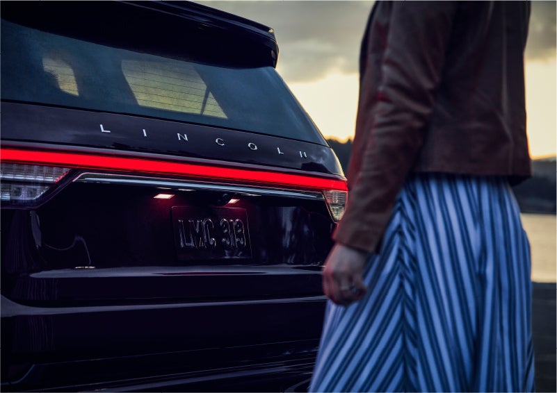 A person is shown near the rear of a 2023 Lincoln Aviator® SUV as the Lincoln Embrace illuminates the rear lights | Biondi Lincoln in Monroeville PA