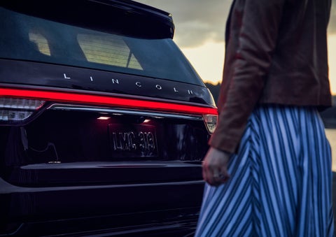 A person is shown near the rear of a 2024 Lincoln Aviator® SUV as the Lincoln Embrace illuminates the rear lights | Biondi Lincoln in Monroeville PA