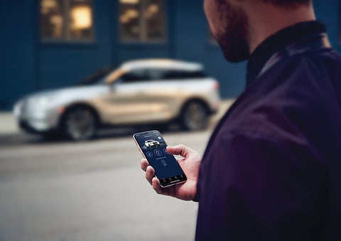 A person is shown interacting with a smartphone to connect to a Lincoln vehicle across the street. | Biondi Lincoln in Monroeville PA