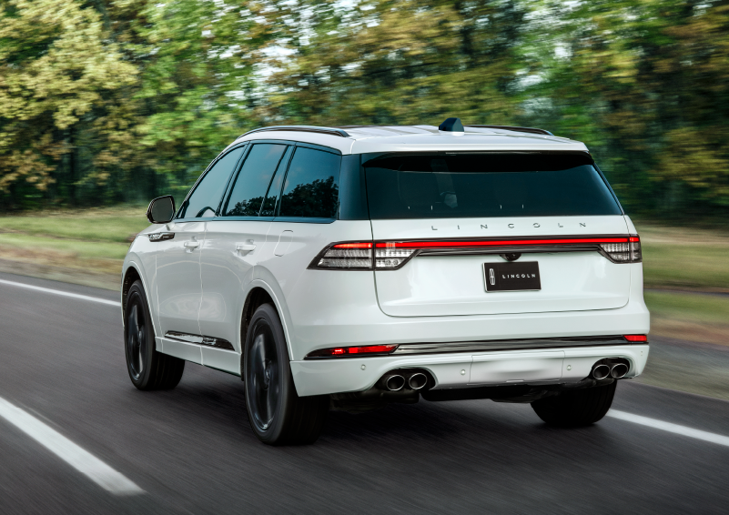 A white 2025 Lincoln Aviator® SUV is being driven away down a road lined with colorful trees. | Biondi Lincoln in Monroeville PA