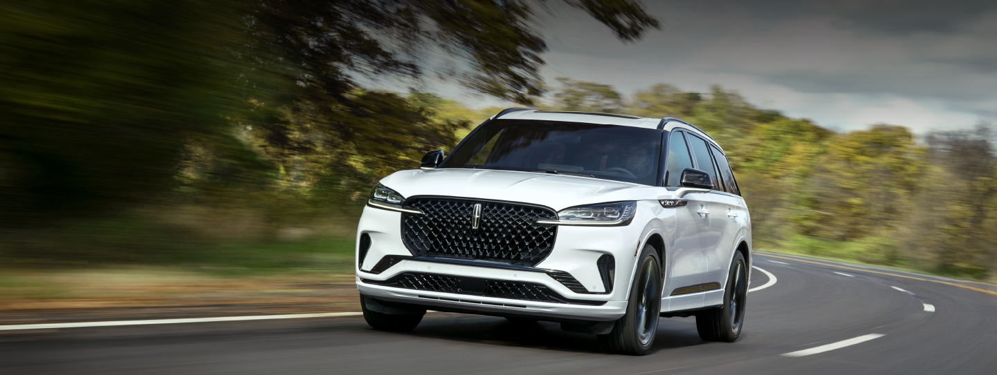 A white 2025 Lincoln Aviator® SUV is being driven down a road lined with trees. | Biondi Lincoln in Monroeville PA