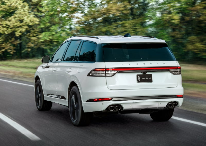A white 2026 Lincoln Aviator® SUV is being driven away down a road lined with colorful trees. | Biondi Lincoln in Monroeville PA
